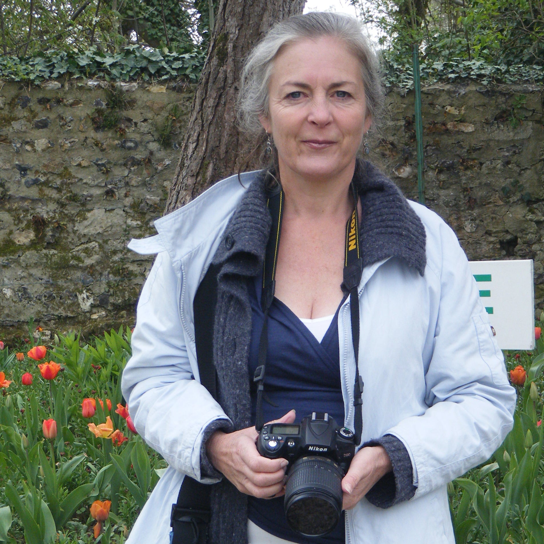 Simone, Yvette's sister,  holding a camera in a garden with tulips and a stone wall in the background